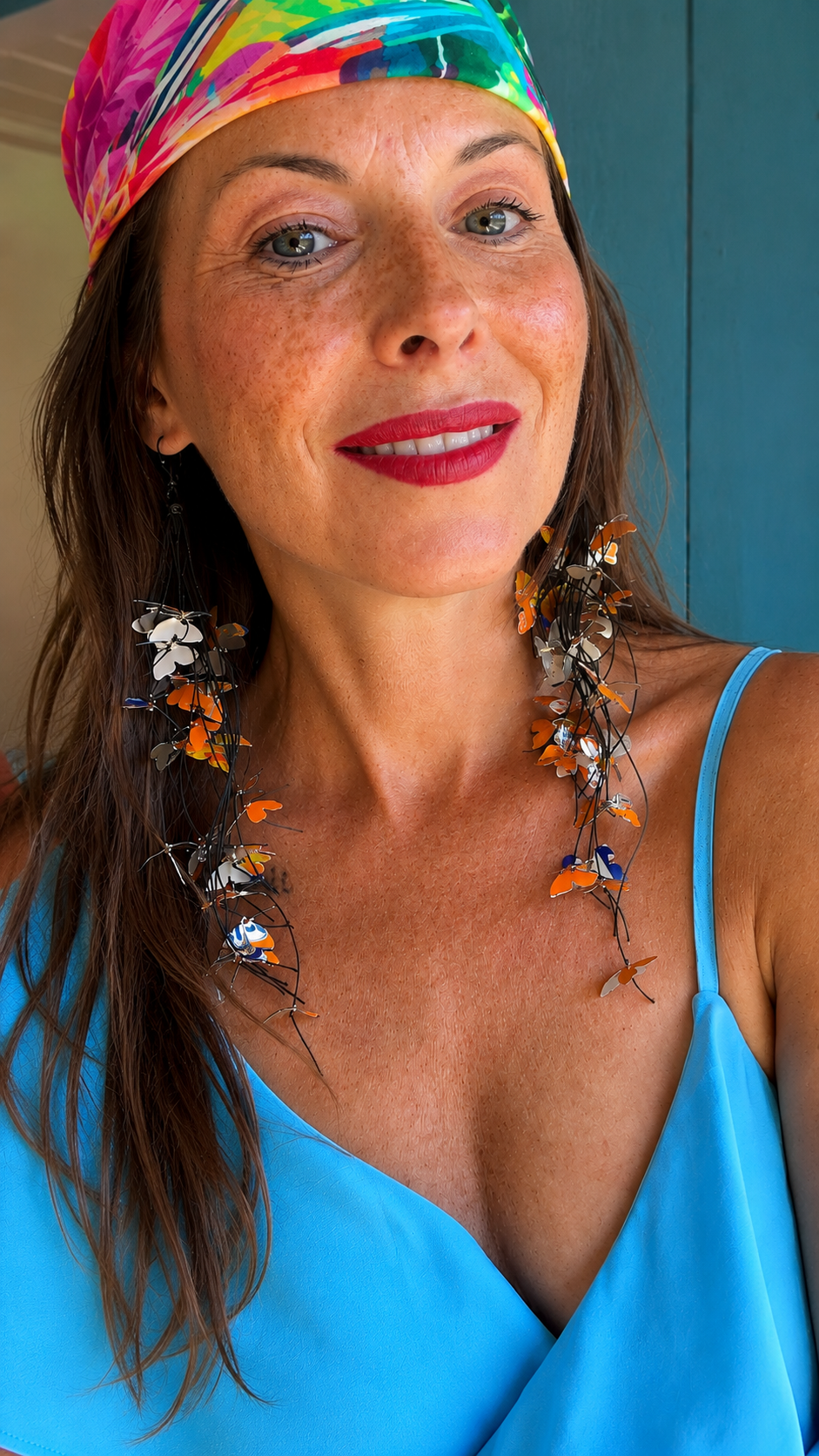 A close-up portrait of a woman wearing a vibrant multicolor butterfly-themed headband and matching dangling earrings made from upcycled fanta cans, paired with a bright blue top.