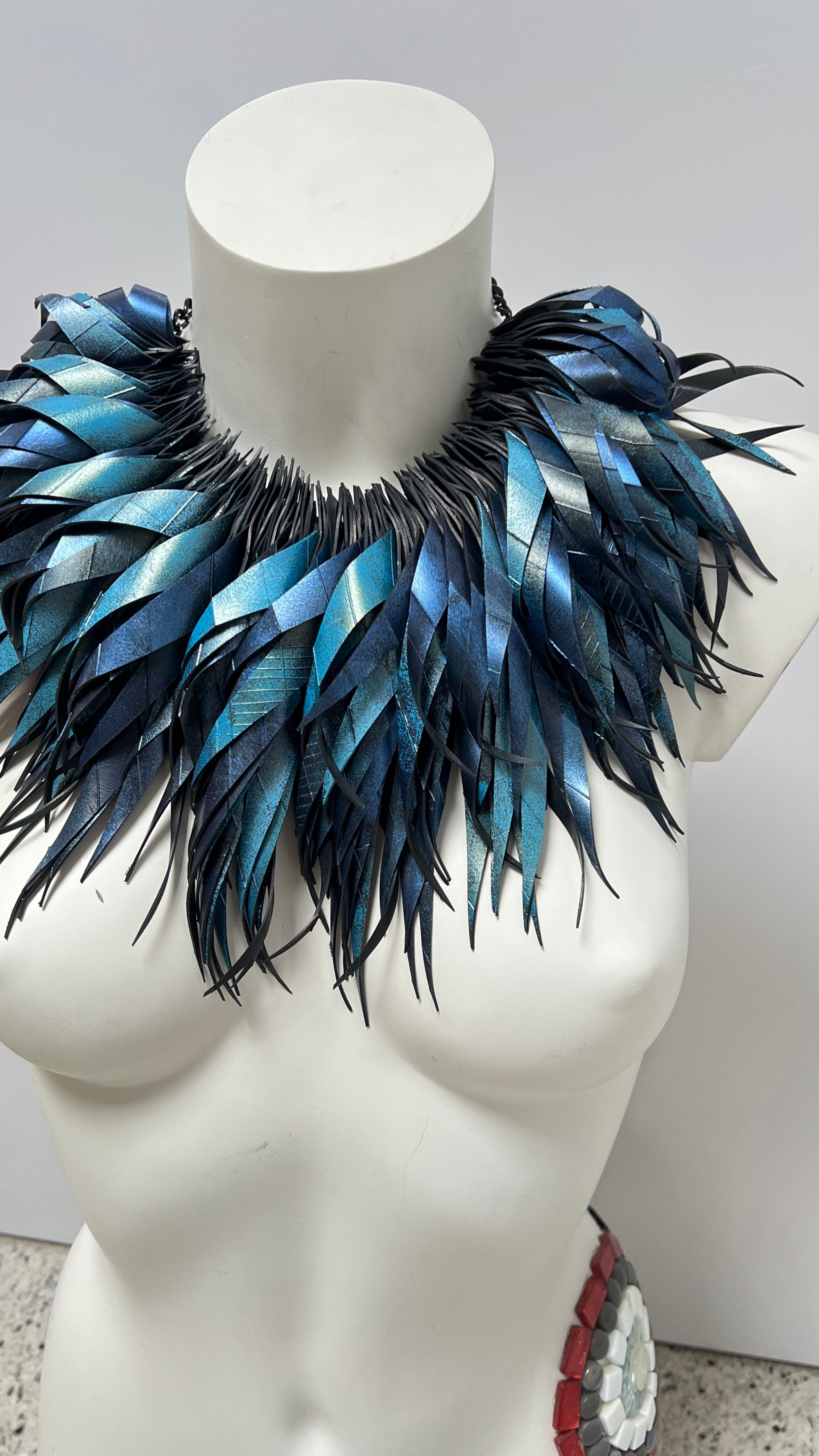 metallic bue coloured large, chunky curly tassel necklace made from upcycled bicycle inner tube on a white mannequin iagainst a white wall