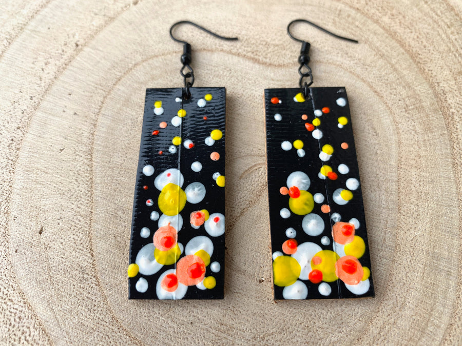 Squared-shaped earrings with black, white, orange, and yellow bubble design, made from upcycled materials on a wooden background