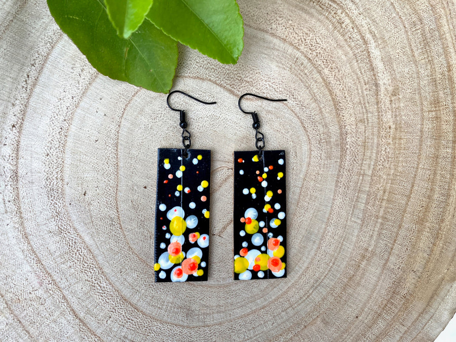 Squared-shaped earrings with black, white, orange, and yellow bubble design, made from upcycled materials on a wooden background