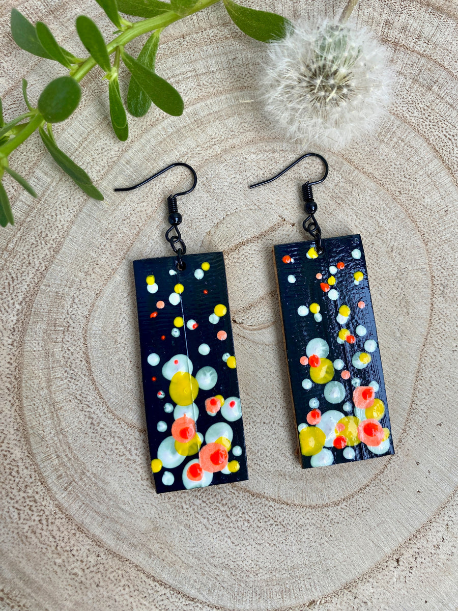 Squared-shaped earrings with black, white, orange, and yellow bubble design, made from upcycled materials on a wooden background