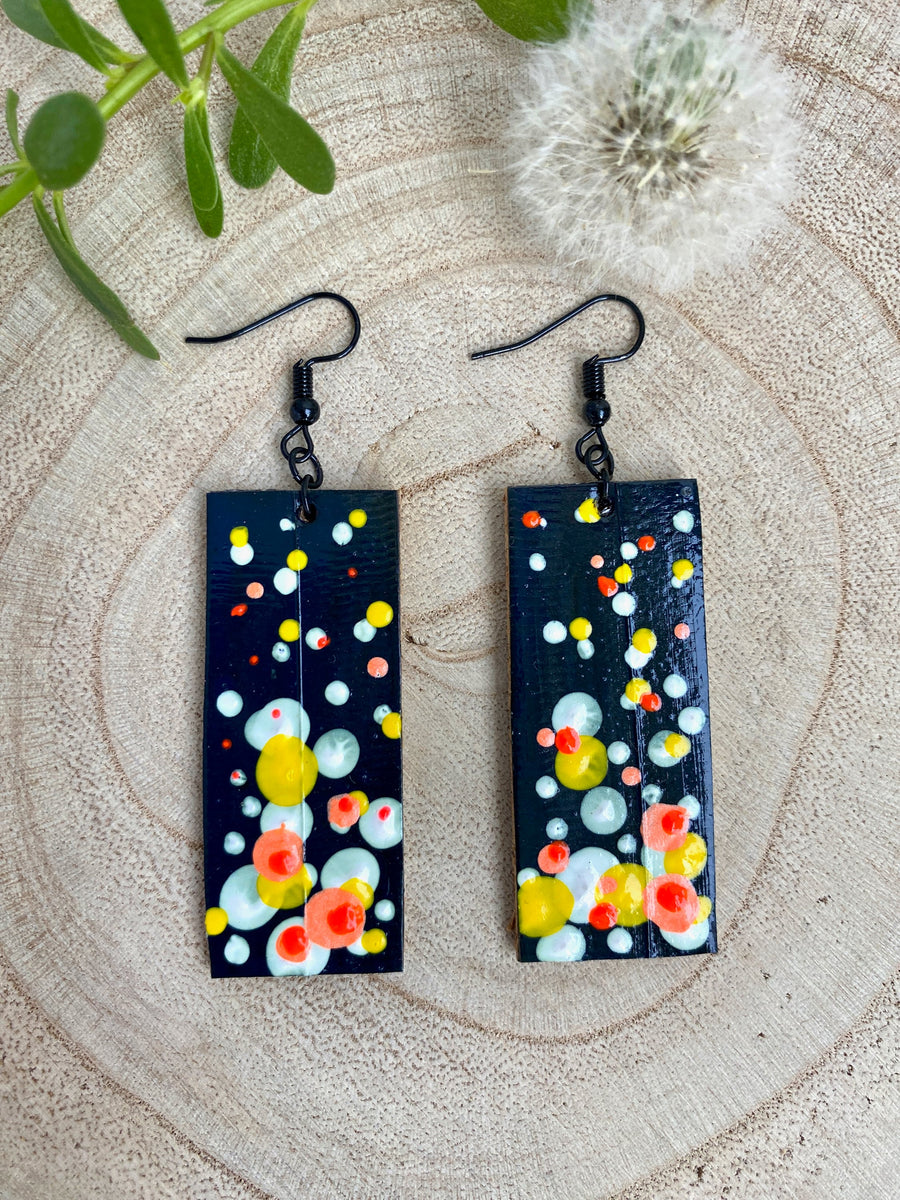 Squared-shaped earrings with black, white, orange, and yellow bubble design, made from upcycled materials on a wooden background