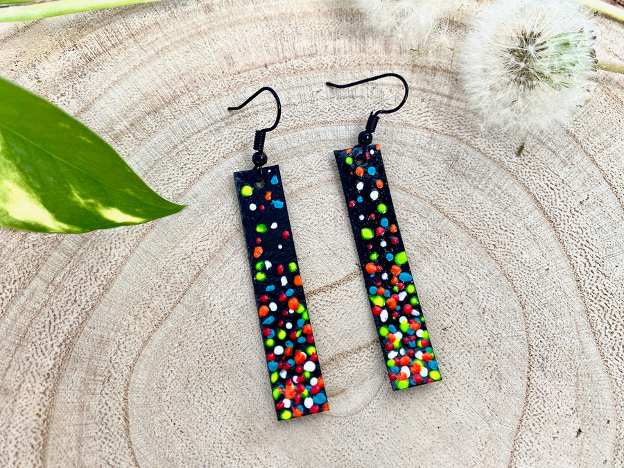 Long, thin black coloured upcycled bicycle inner tube earrings decorated with colourful polka dots on a wooden background with a leaf and dandelions