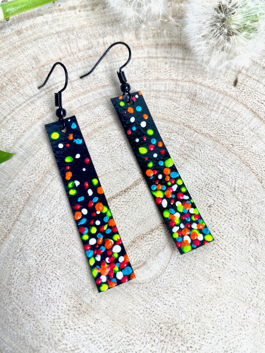 Long, thin black coloured upcycled bicycle inner tube earrings decorated with colourful polka dots on a wooden background with a leaf and dandelions