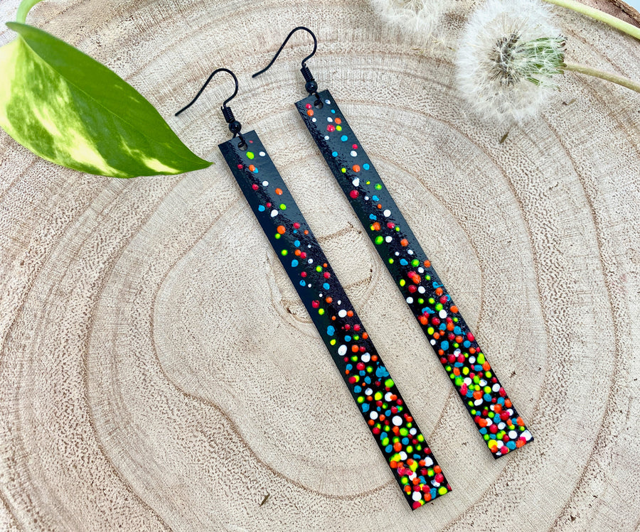 Long, black upcycled inner tube strap shaped earrings decorated with colourful polka dots, on a wooden background with dandelions