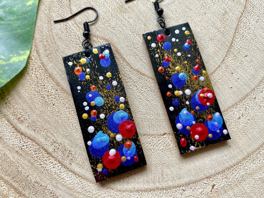 squared, black earrings with blue, red, golden and silver polka dots on a wooden background with a leaf