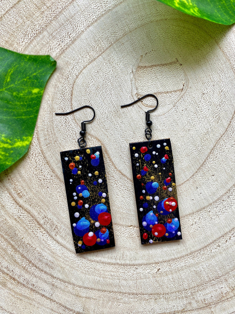 squared, black earrings with blue, red, golden and silver polka dots on a wooden background with a leaf