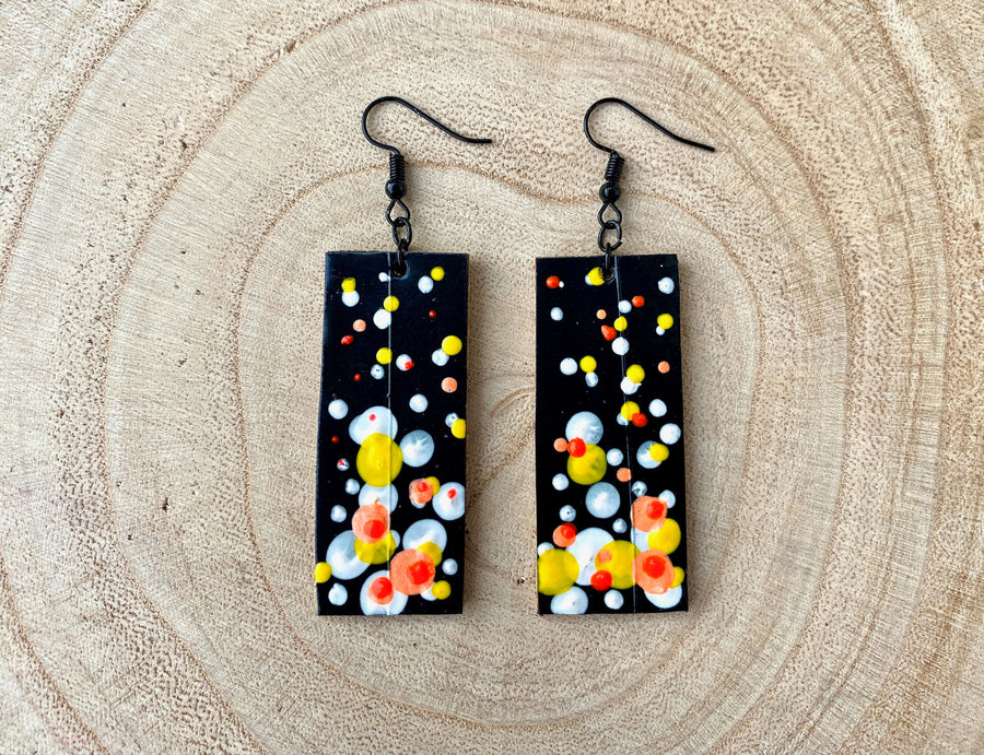 Squared-shaped earrings with black, white, orange, and yellow bubble design, made from upcycled materials on a wooden background