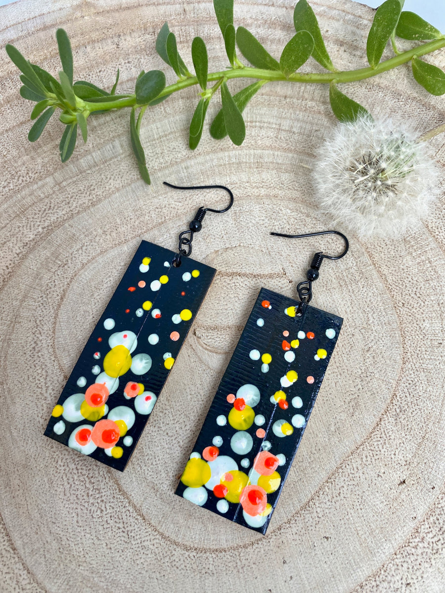 Squared-shaped earrings with black, white, orange, and yellow bubble design, made from upcycled materials on a wooden background