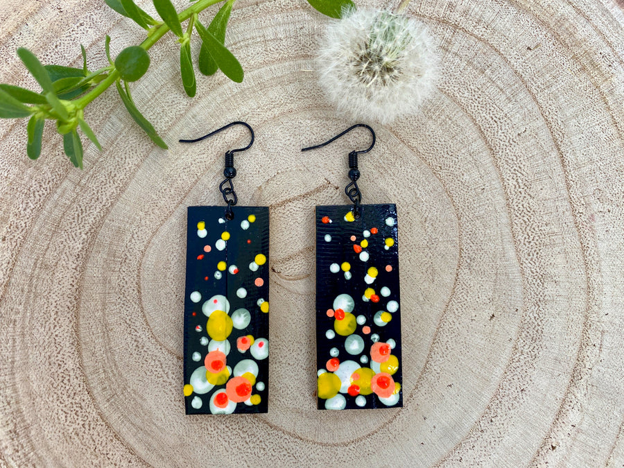 Squared-shaped earrings with black, white, orange, and yellow bubble design, made from upcycled materials on a wooden background