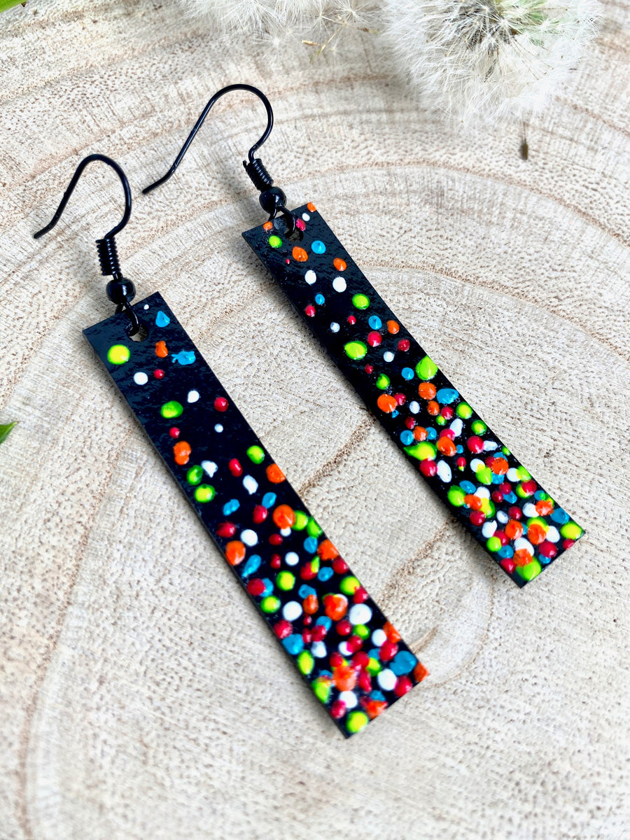 Long, thin black coloured upcycled bicycle inner tube earrings decorated with colourful polka dots on a wooden background with a leaf and dandelions