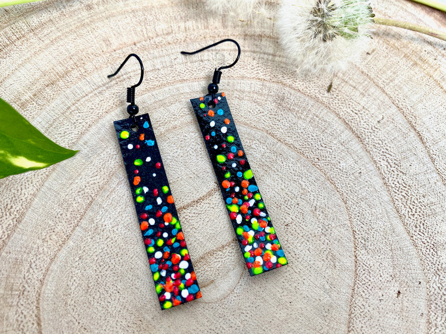 Long, thin black coloured upcycled bicycle inner tube earrings decorated with colourful polka dots on a wooden background with a leaf and dandelions