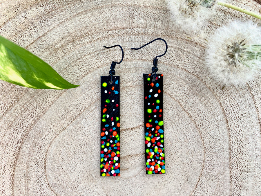 Long, thin black coloured upcycled bicycle inner tube earrings decorated with colourful polka dots on a wooden background with a leaf and dandelions