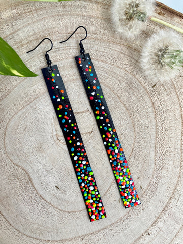 Long, black upcycled inner tube strap shaped earrings decorated with colourful polka dots, on a wooden background with dandelions