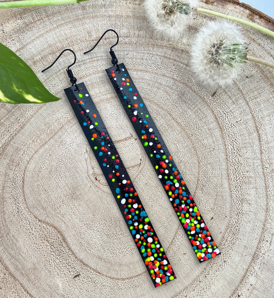 Long, black upcycled inner tube strap shaped earrings decorated with colourful polka dots, on a wooden background with dandelions