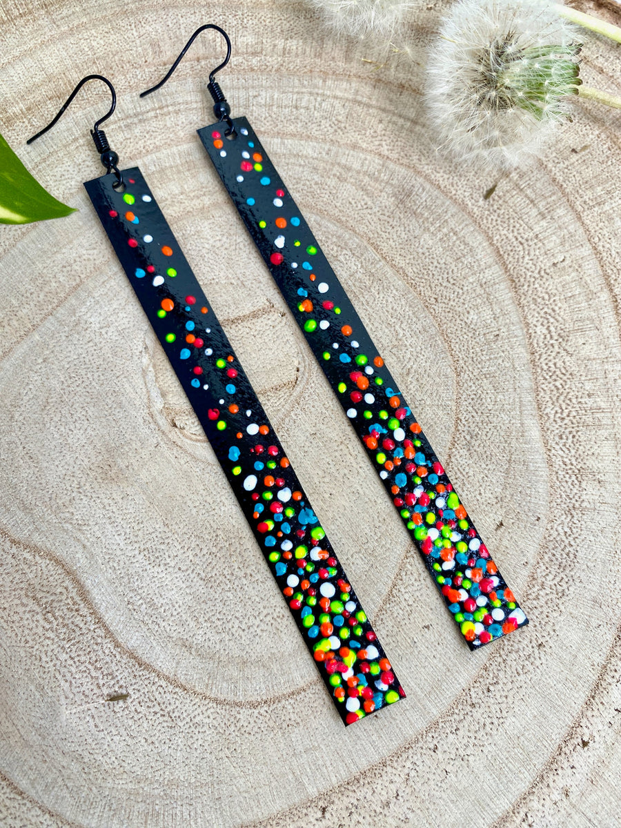 Long, black upcycled inner tube strap shaped earrings decorated with colourful polka dots, on a wooden background with dandelions