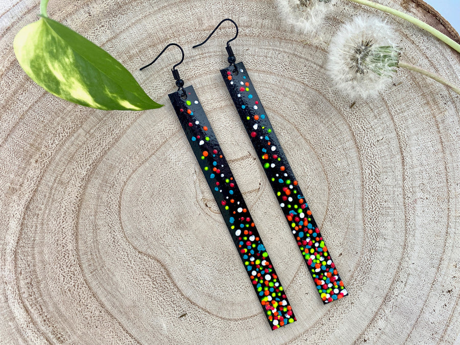 Long, black upcycled inner tube strap shaped earrings decorated with colourful polka dots, on a wooden background with dandelions