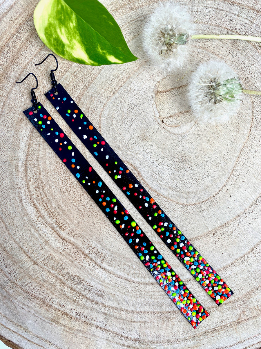 Long, black upcycled bike inner tube earrings decorated with colourful polka dots on a wooden background with 2 dandelions