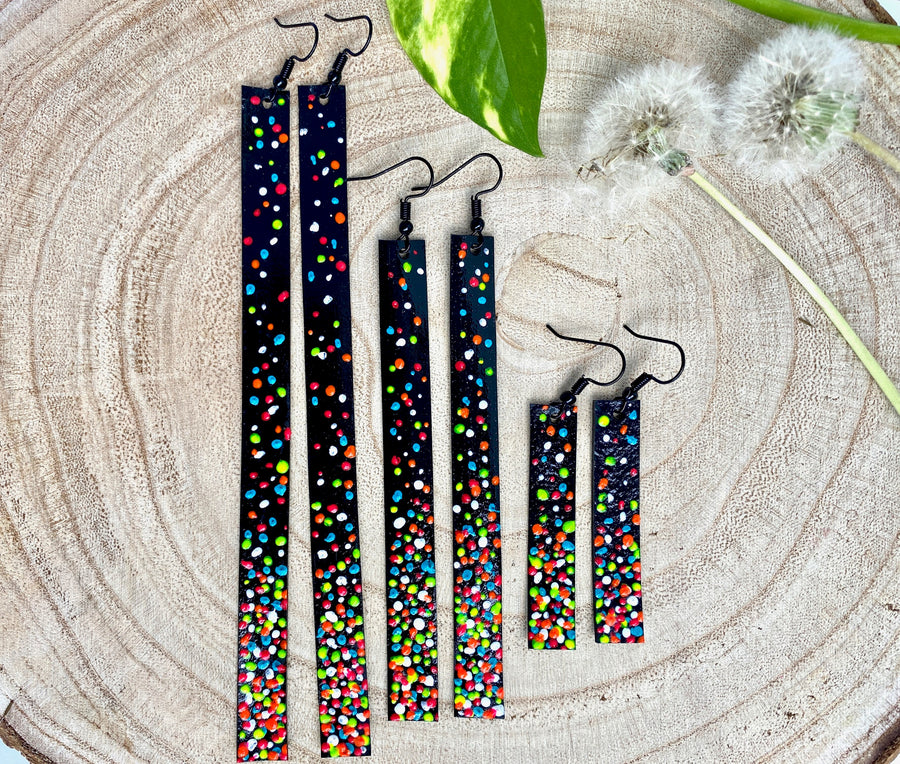 3 pairs of black upcycled bike inner tube earrings decorated with colourful polka dots on a wooden background with 2 dandelions