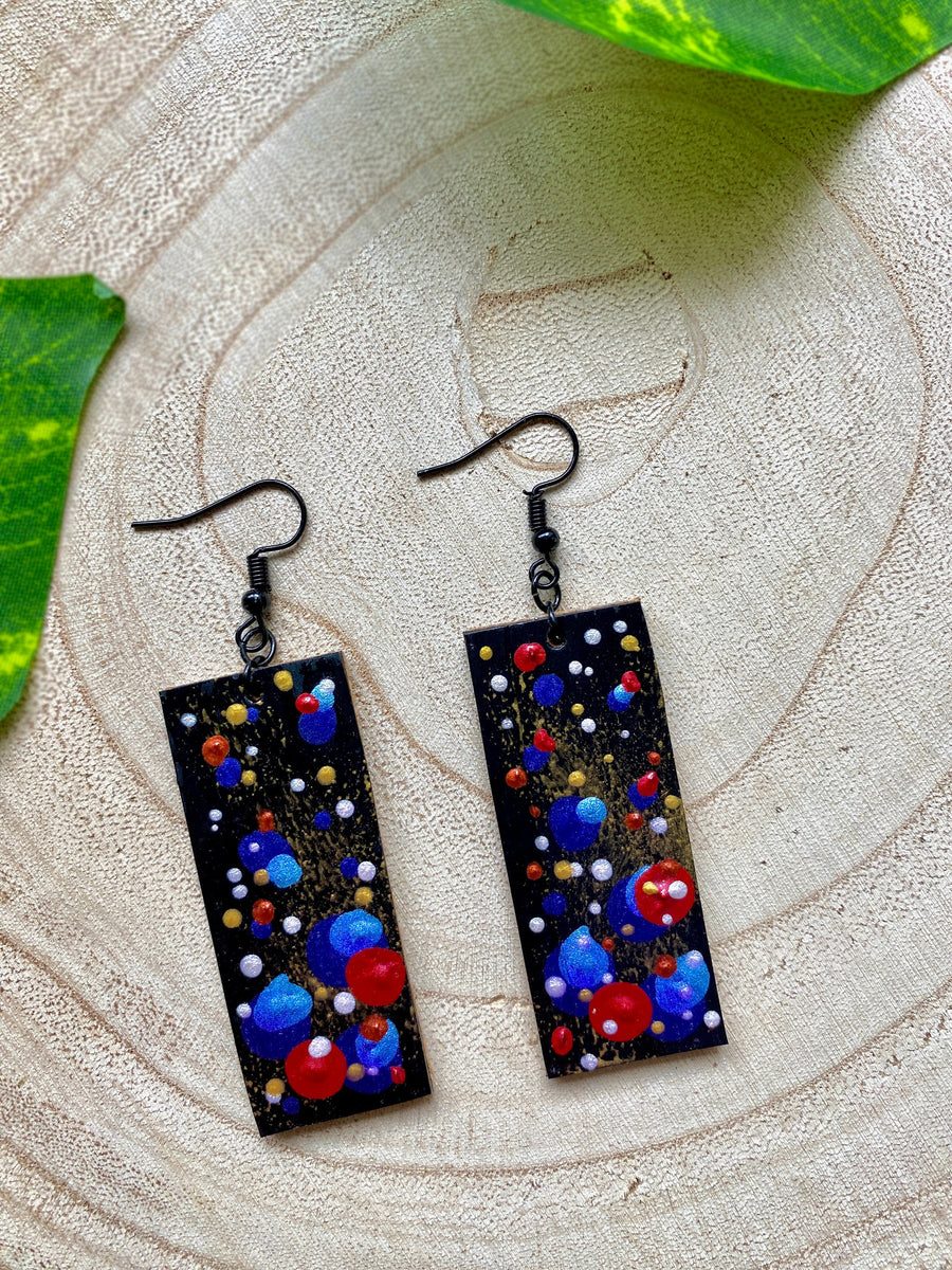 squared, black earrings with blue, red, golden and silver polka dots on a wooden background with a leaf