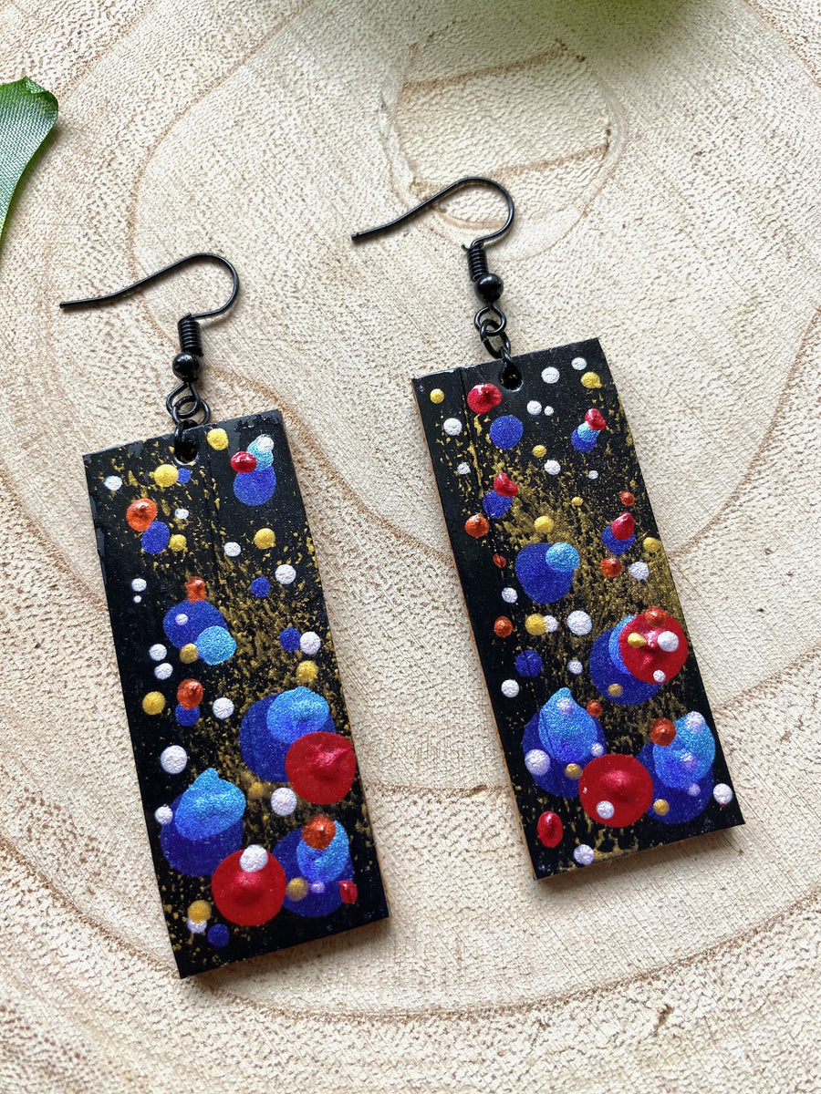 squared, black earrings with blue, red, golden and silver polka dots on a wooden background with a leaf