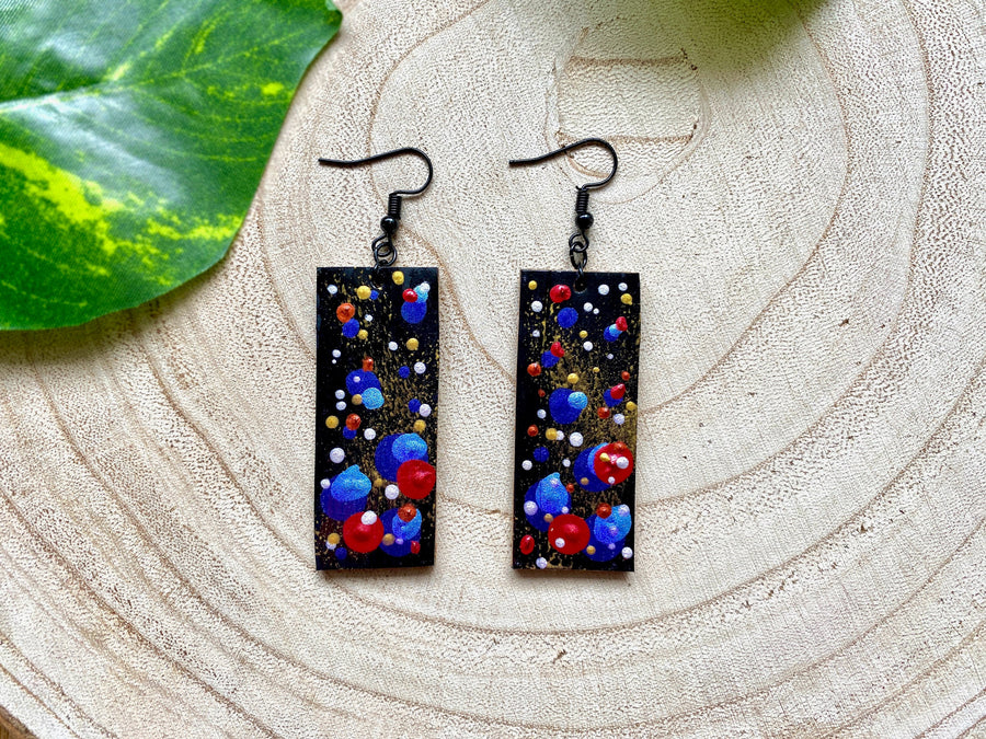 squared, black earrings with blue, red, golden and silver polka dots on a wooden background with a leaf