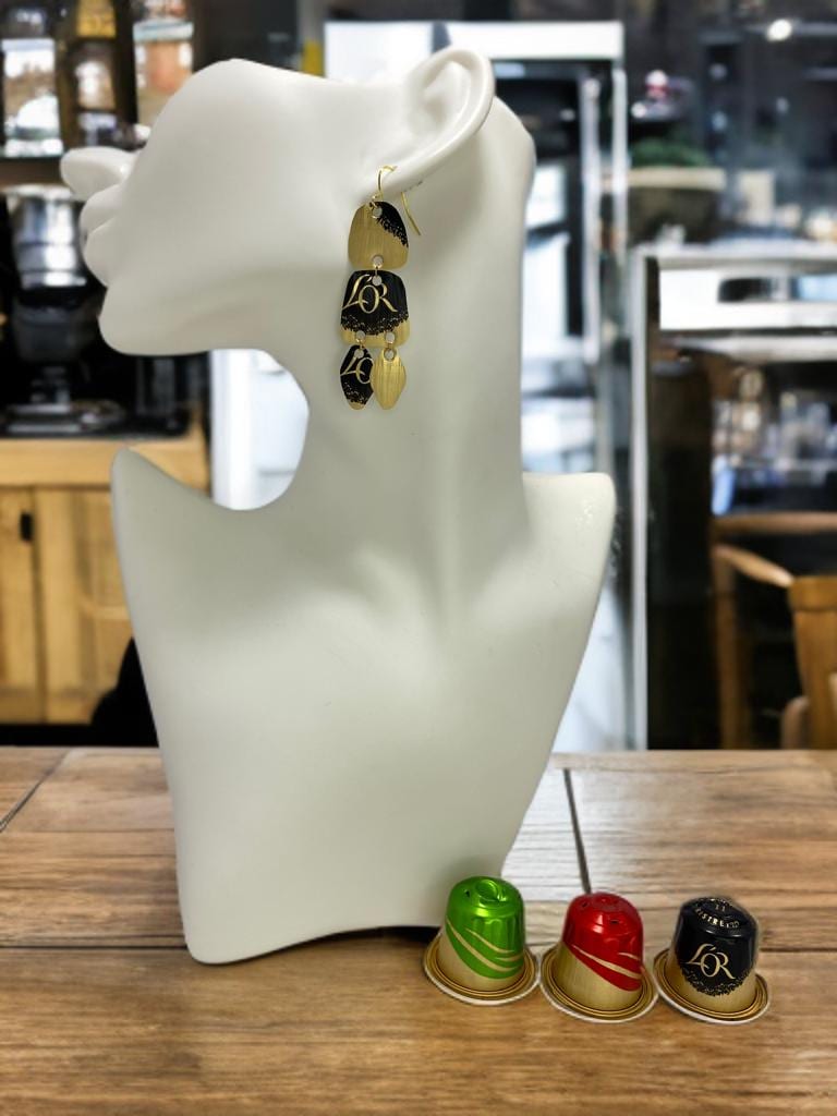 Upcycled black-golden Nespresso capsules earrings on a white mannequin display with 3 nespresso pods on a bar desk