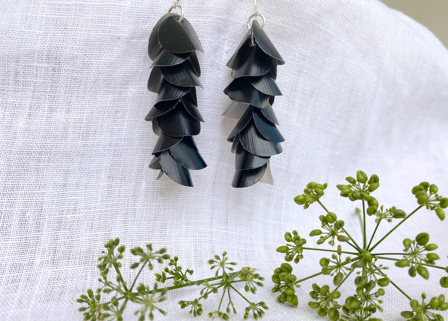 a pair of black, cascading aluminium earrings made from upcycled nespresso capsules on a white linen background and green flowers