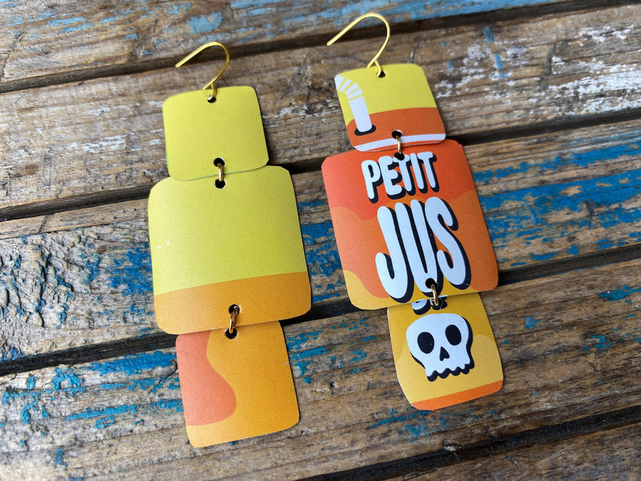 pair of yellow orange, asymmetric geometric earrings with text and skull design made from an upcycled beer can on a rustic wooden background