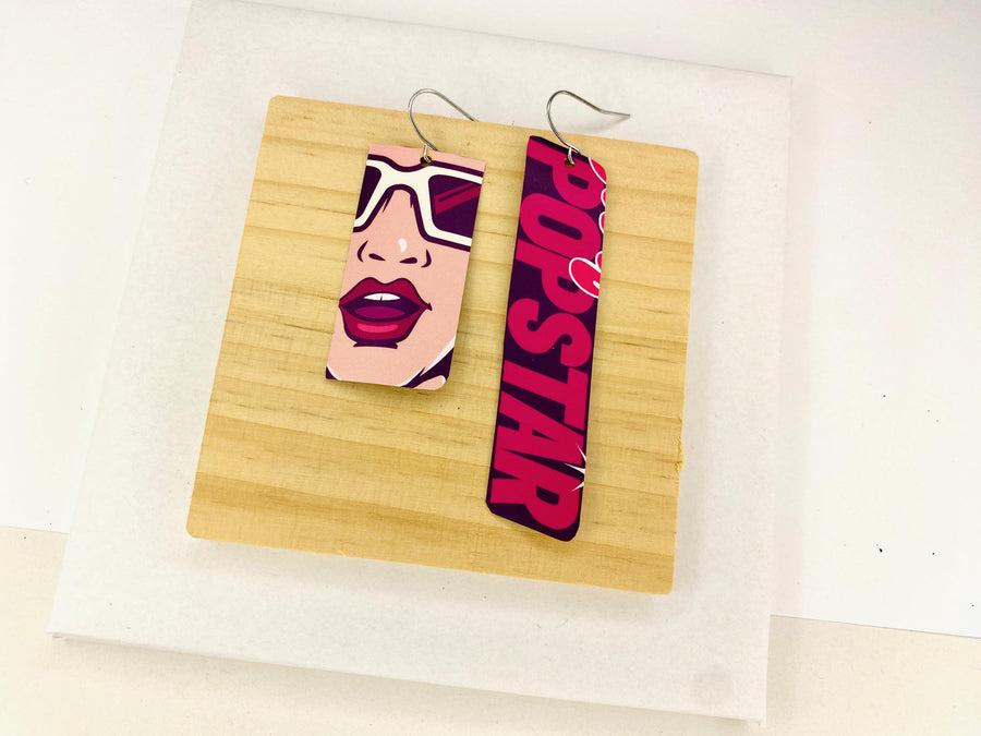 asymmetric, 90's Pop-art earrings featuring POPSTAR text and women's face made from  an upcycled craft beer can on a wooden and white background