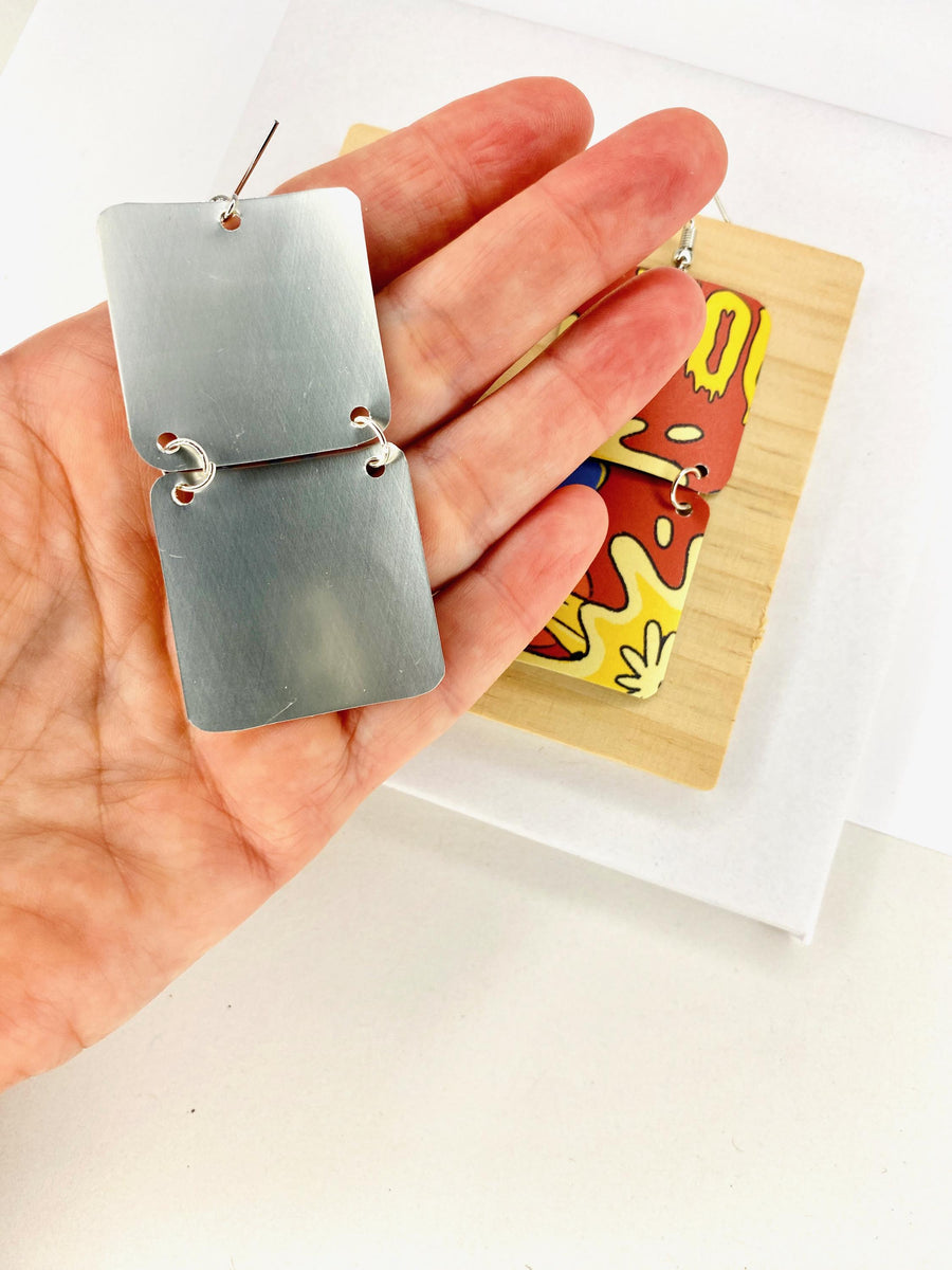 comic, graffiti style earrings made from an upcycled craft beer can in orange, yellow colours, on a wooden and white background and displayed in hand