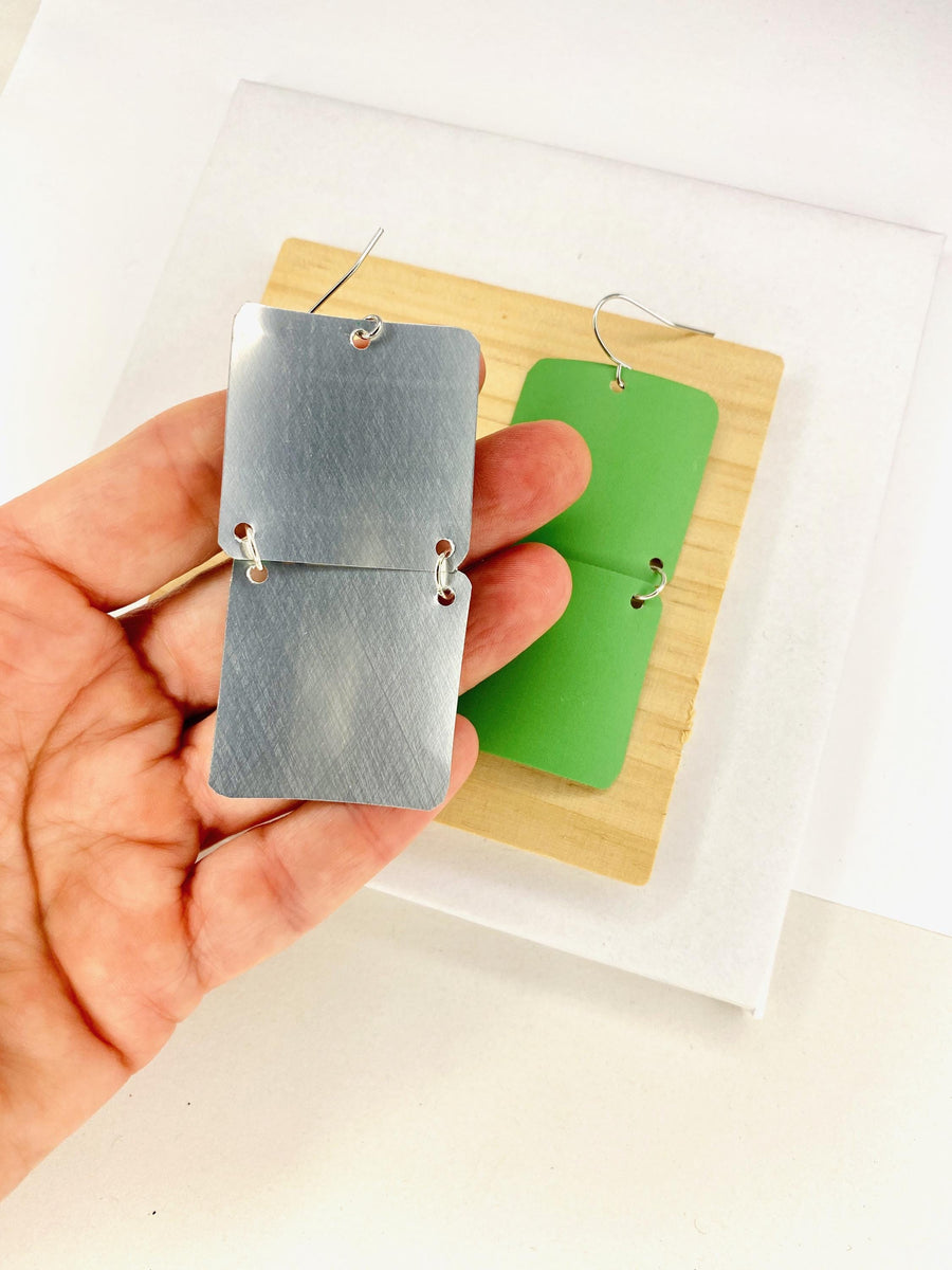 green earrings made from a craft beer can from Other Half Brewery on a wooden and white background and displayed in hand