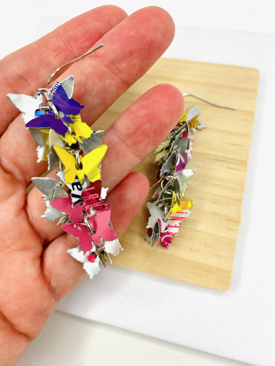 colourful, cascade statement earrings featuring tiny butterflies that are upcycled from craft beer cans on a wooden and white background