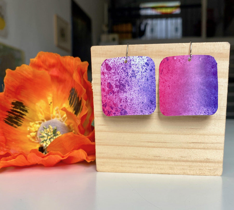 pink purple sqaured pop-art earrings with milky way design made from upcycled craft beer cans, Laura zabo design, on a wooden background and flower decoration