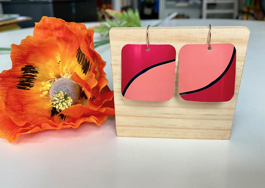 Laura Zabo , dark and light pink squared earrings made from shiny craft beer can, on a wooden background and flower decoration
