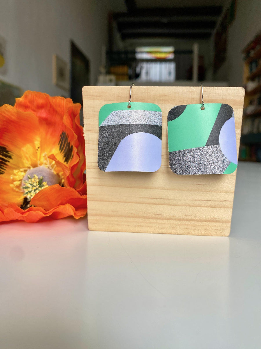 grey green coloured squared geometric earrings made from upcycled craft beer cans by laura zabo on a wooden background and flower