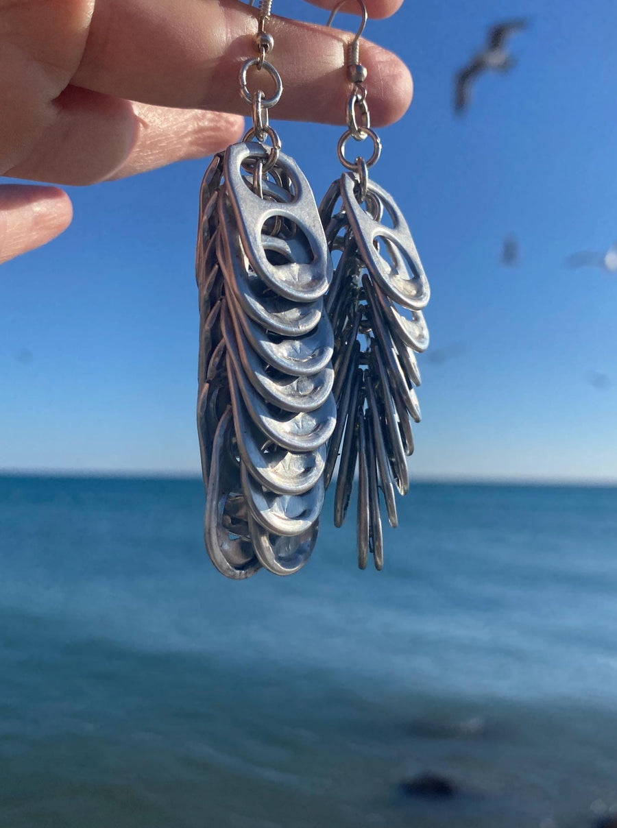Silver coloured statement earrings made from Craft Beer cans hang on fingers near the oceans and birds