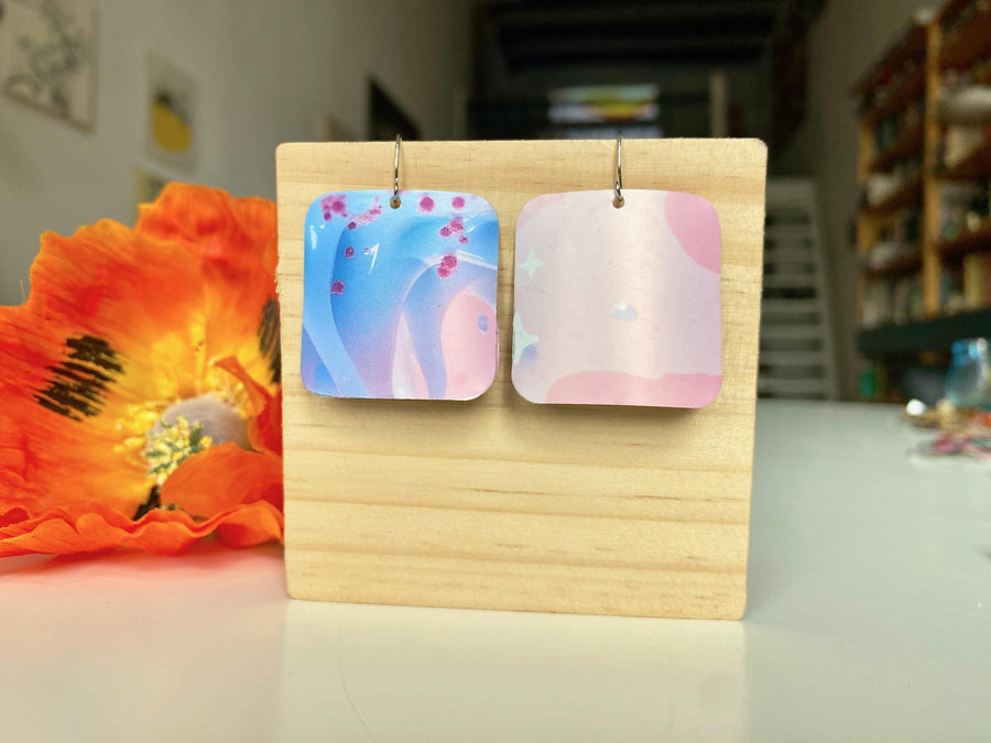 light blue and pink, dreamy, squared earrings made from upcycled craft beer can by laura zabo on a wooden display and orange flower