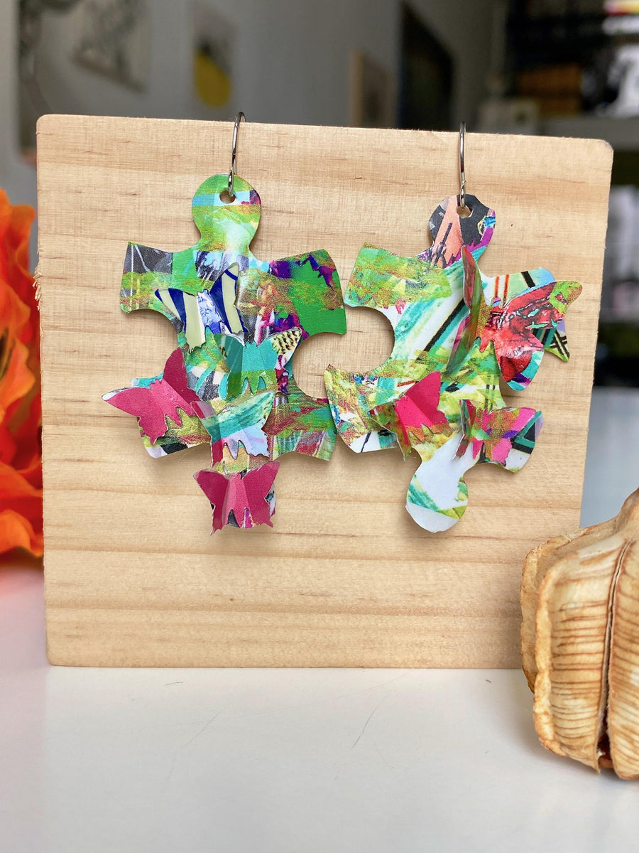 colourful, puzzle shaped earrings adorned with tiny butterflies made from upcycled craft beer cans by laura zabo on a wooden background and flower design