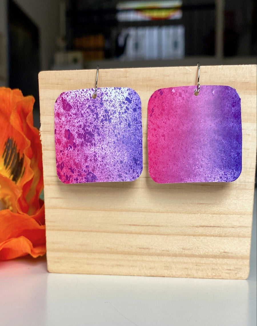 pink purple sqaured pop-art earrings with milky way design made from upcycled craft beer cans, Laura zabo design, on a wooden background and flower decoration