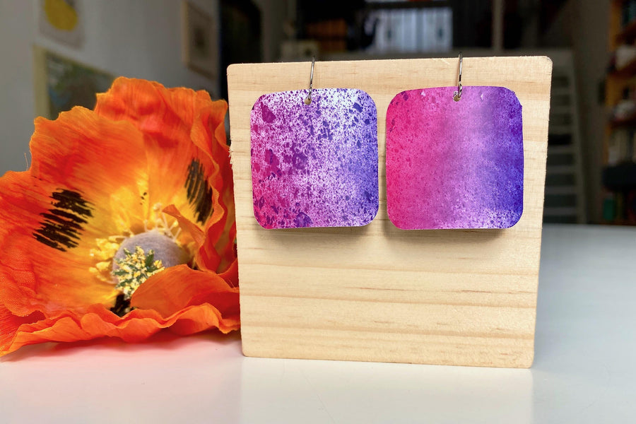 pink purple sqaured pop-art earrings with milky way design made from upcycled craft beer cans, Laura zabo design, on a wooden background and flower decoration