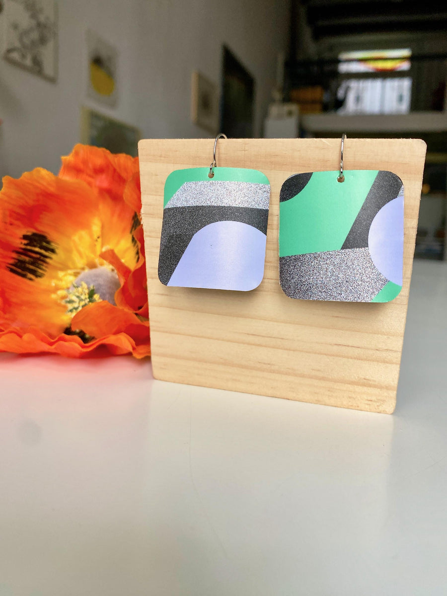 grey green coloured squared geometric earrings made from upcycled craft beer cans by laura zabo on a wooden background and flower