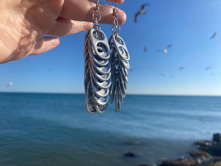 Silver coloured statement earrings made from Craft Beer cans hang on fingers near the oceans and birds