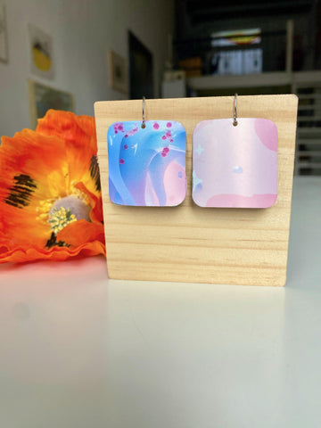 light blue and pink, dreamy, squared earrings made from upcycled craft beer can by laura zabo on a wooden display and orange flower