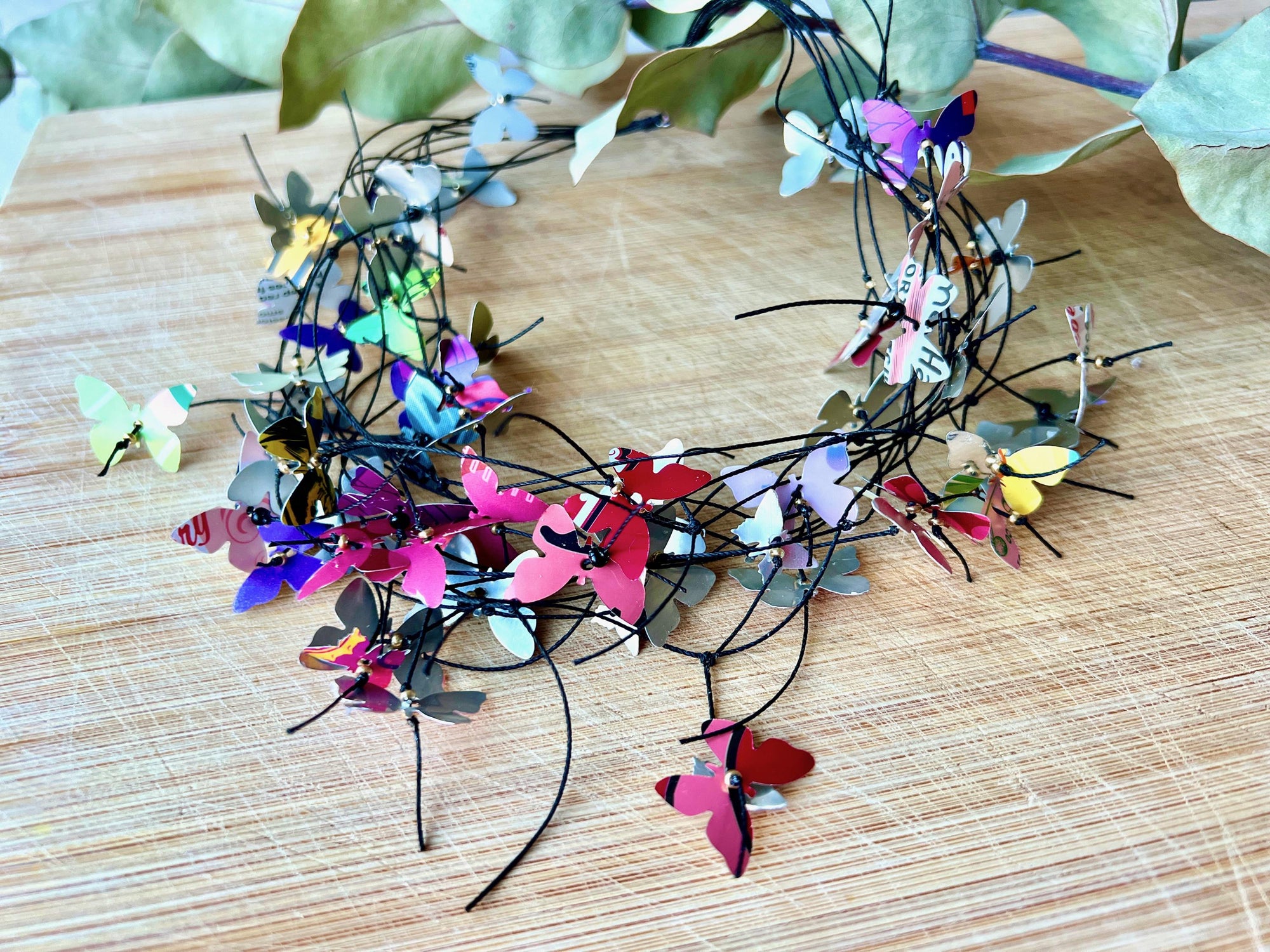elegant, colourful necklace featuring tiny aluminium butterflies, each one is cut from soda and beer cans, fitted on multiple thin black waxed cotton cord on a wooden background