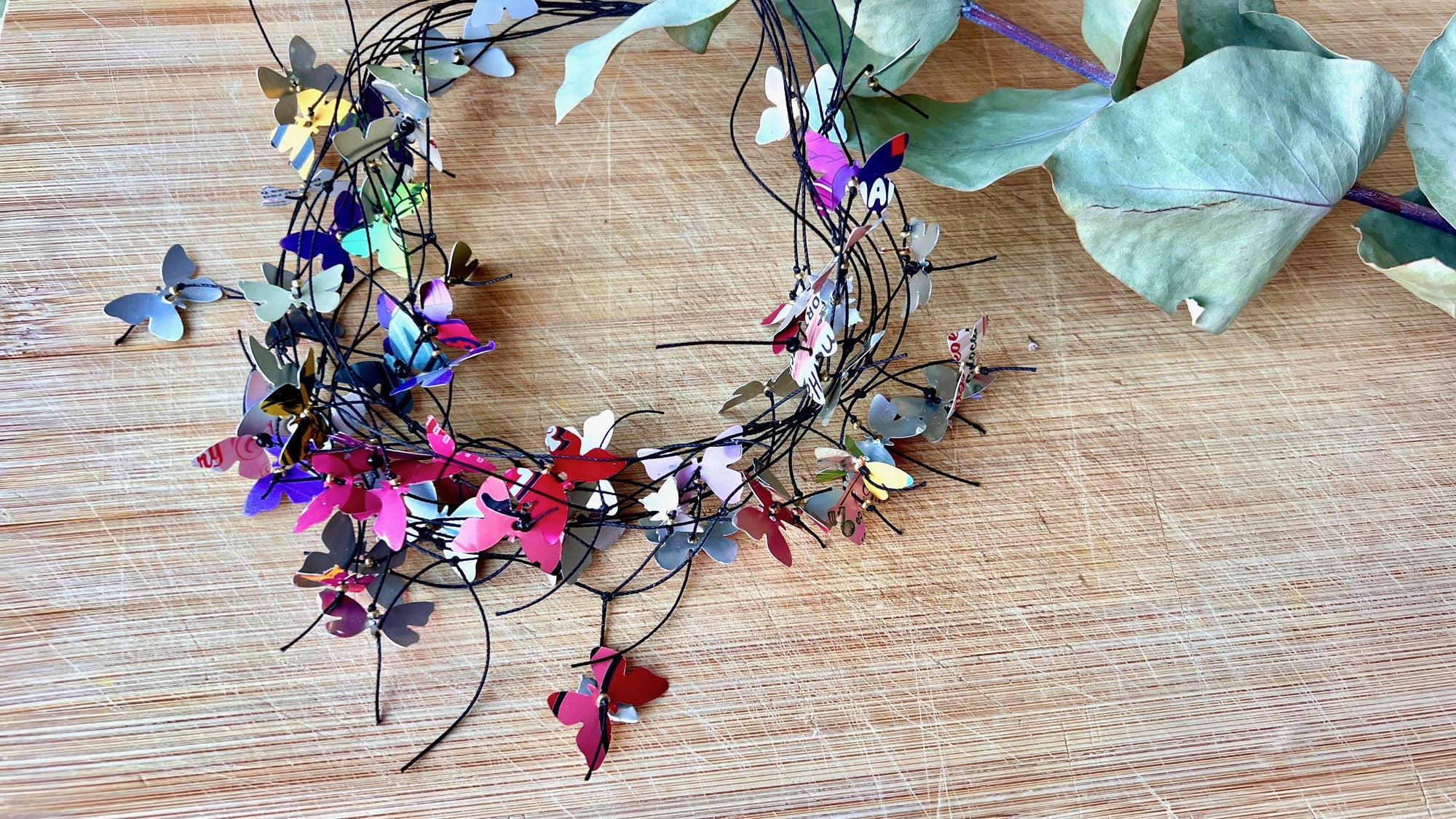 elegant, colourful necklace featuring tiny aluminium butterflies, each one is cut from soda and beer cans, fitted on multiple thin black waxed cotton cord on a wooden background