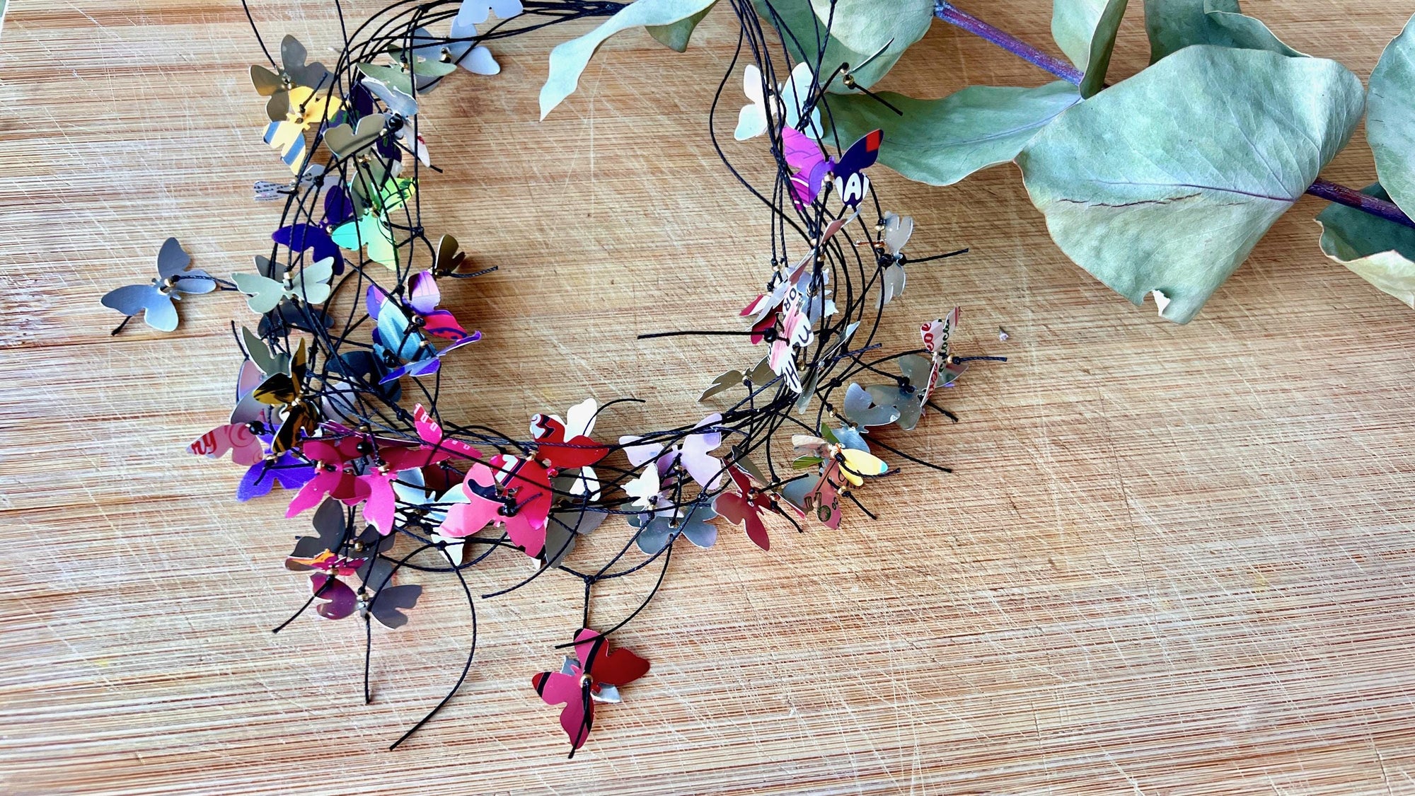 elegant, colourful necklace featuring tiny aluminium butterflies, each one is cut from soda and beer cans, fitted on multiple thin black waxed cotton cord on a wooden background