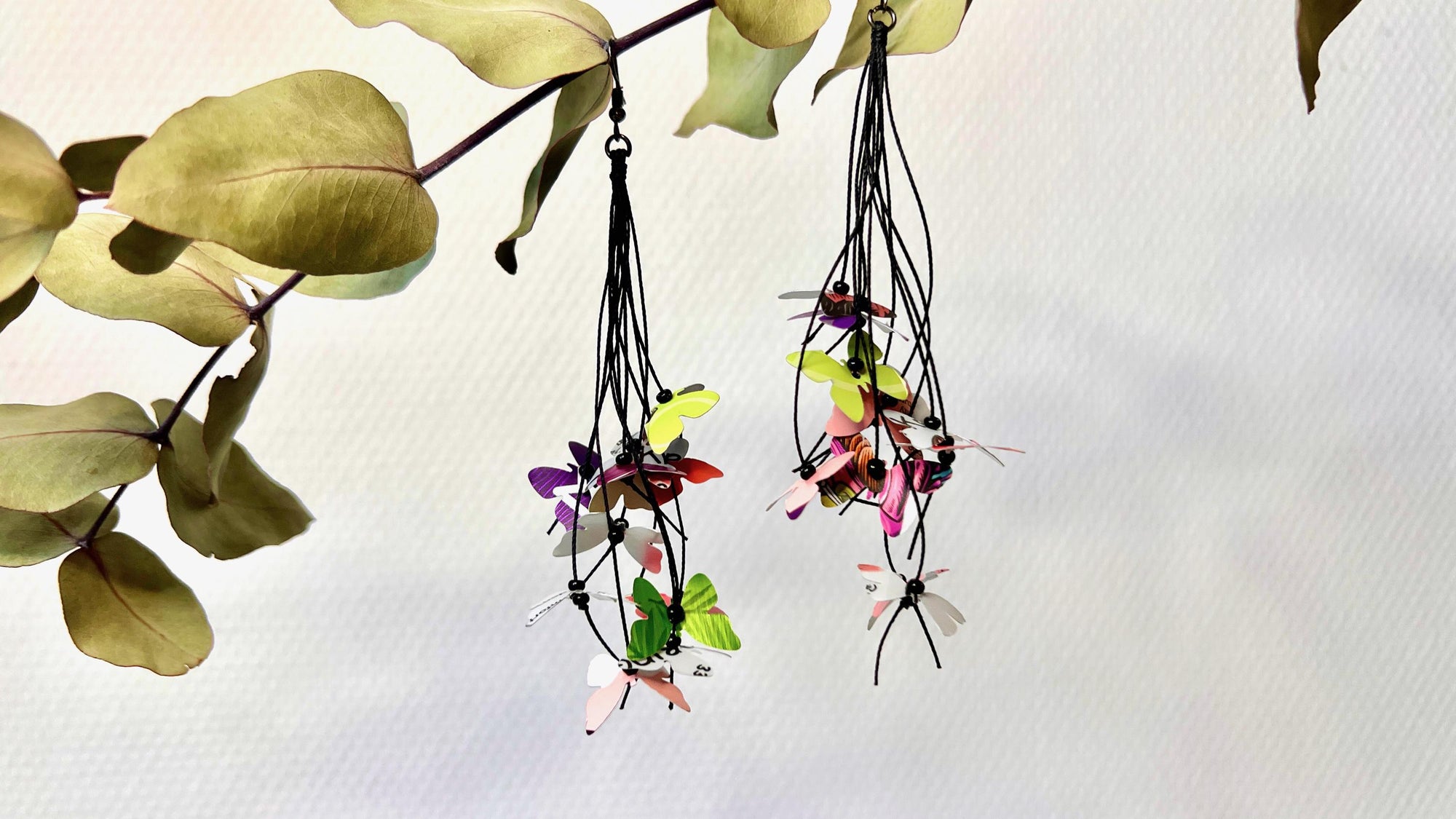 Upcycled earrings hanging on a dry wooden stick with leaves, the earrings feature tiny butterflies cut from soda and beer cans, fitted on waxed cotton cord