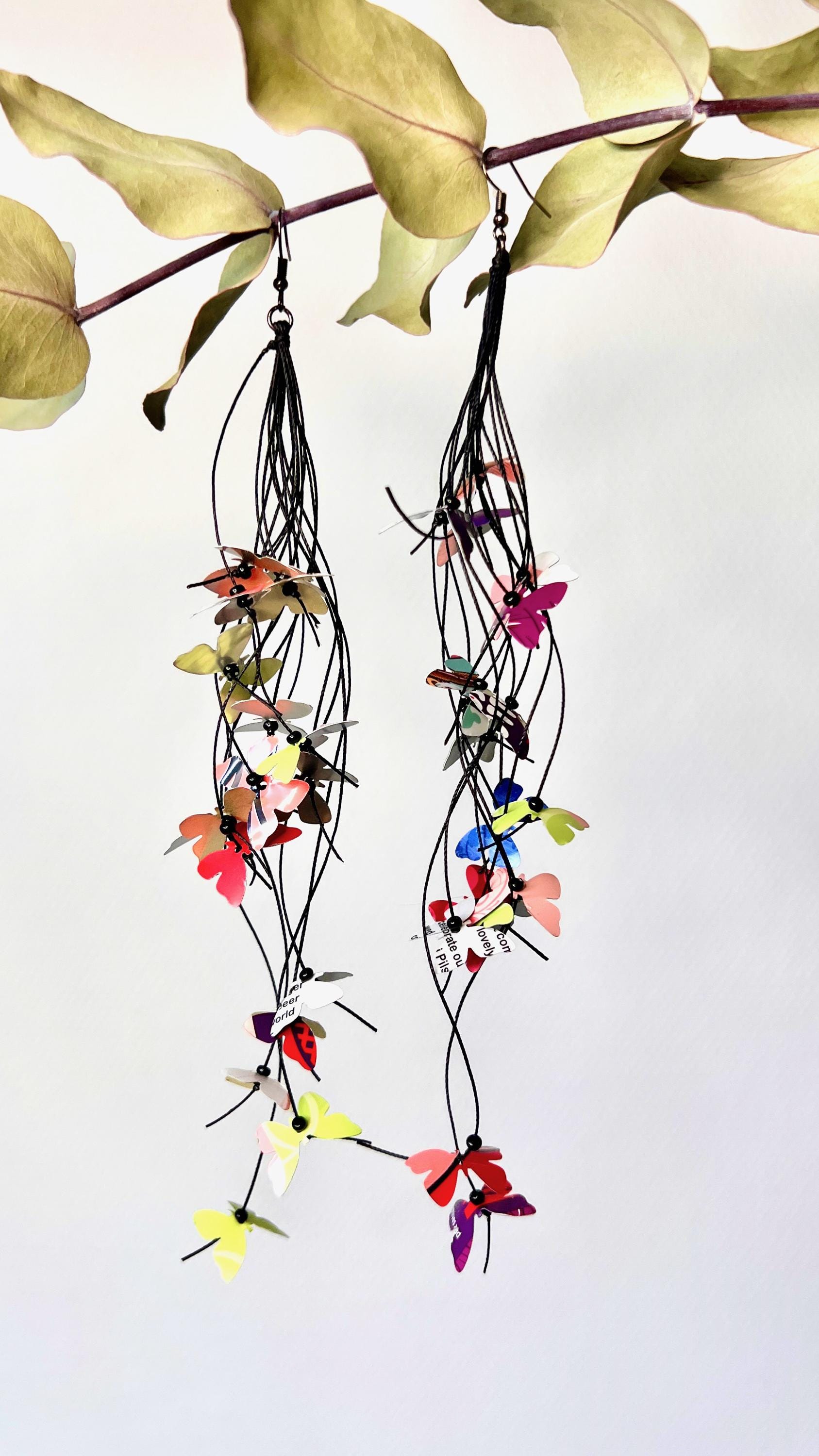 extra long, upcycled earrings hanging on a dry wooden stick with leaves, the earrings feature tiny butterflies cut from soda and beer cans, fitted on waxed cotton cord
