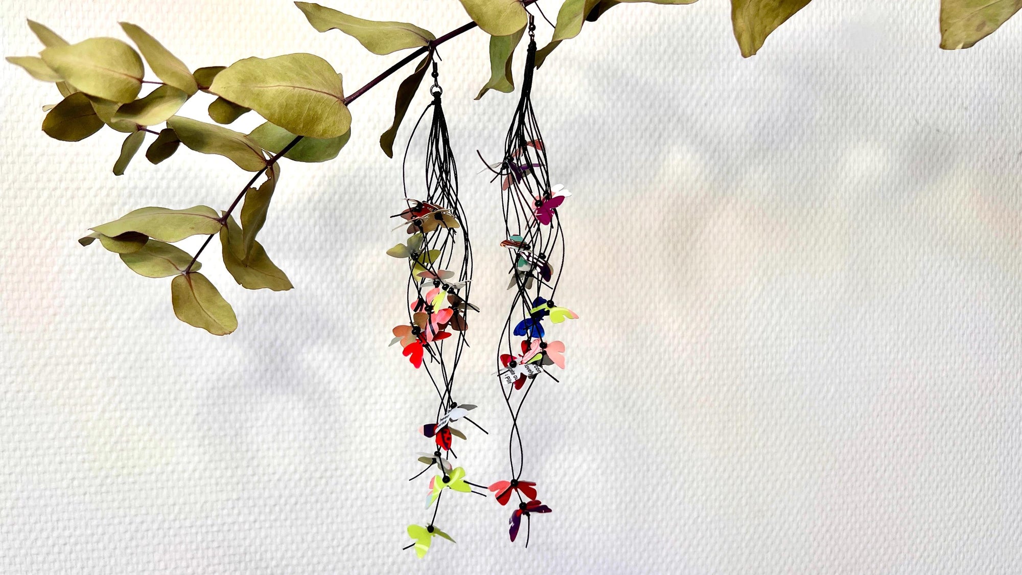extra long, upcycled earrings hanging on a dry wooden stick with leaves, the earrings feature tiny butterflies cut from soda and beer cans, fitted on waxed cotton cord